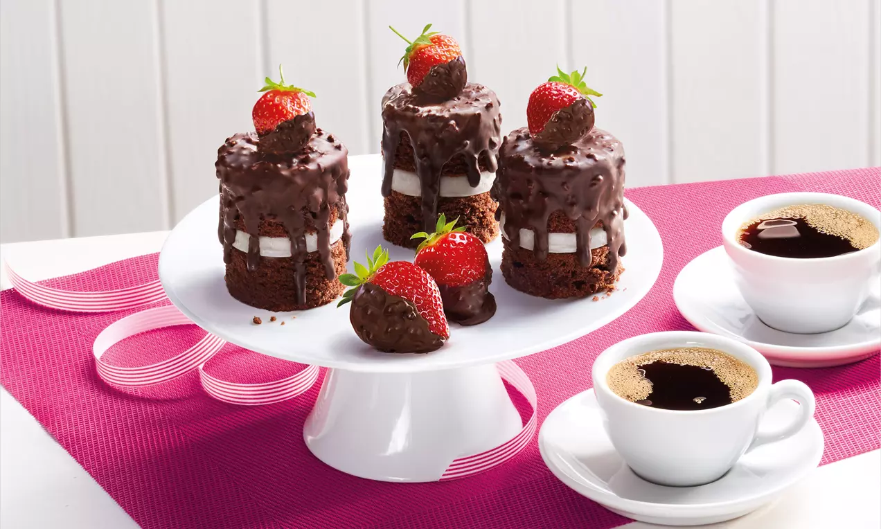 Torrette di torta al cioccolato ricoperte di glassa croccante, decorate con fragole fresche e servite su un piatto bianco accanto a tazze di caffè