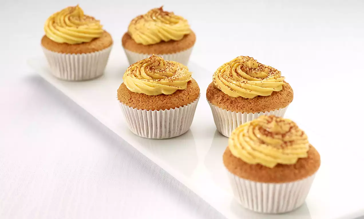 Glutenfreie Baileys-Cupcakes in weißen Papierförmchen, mit goldbeiger Cremehaube und Kakaopulver auf länglicher, weißer Platte angerichtet
