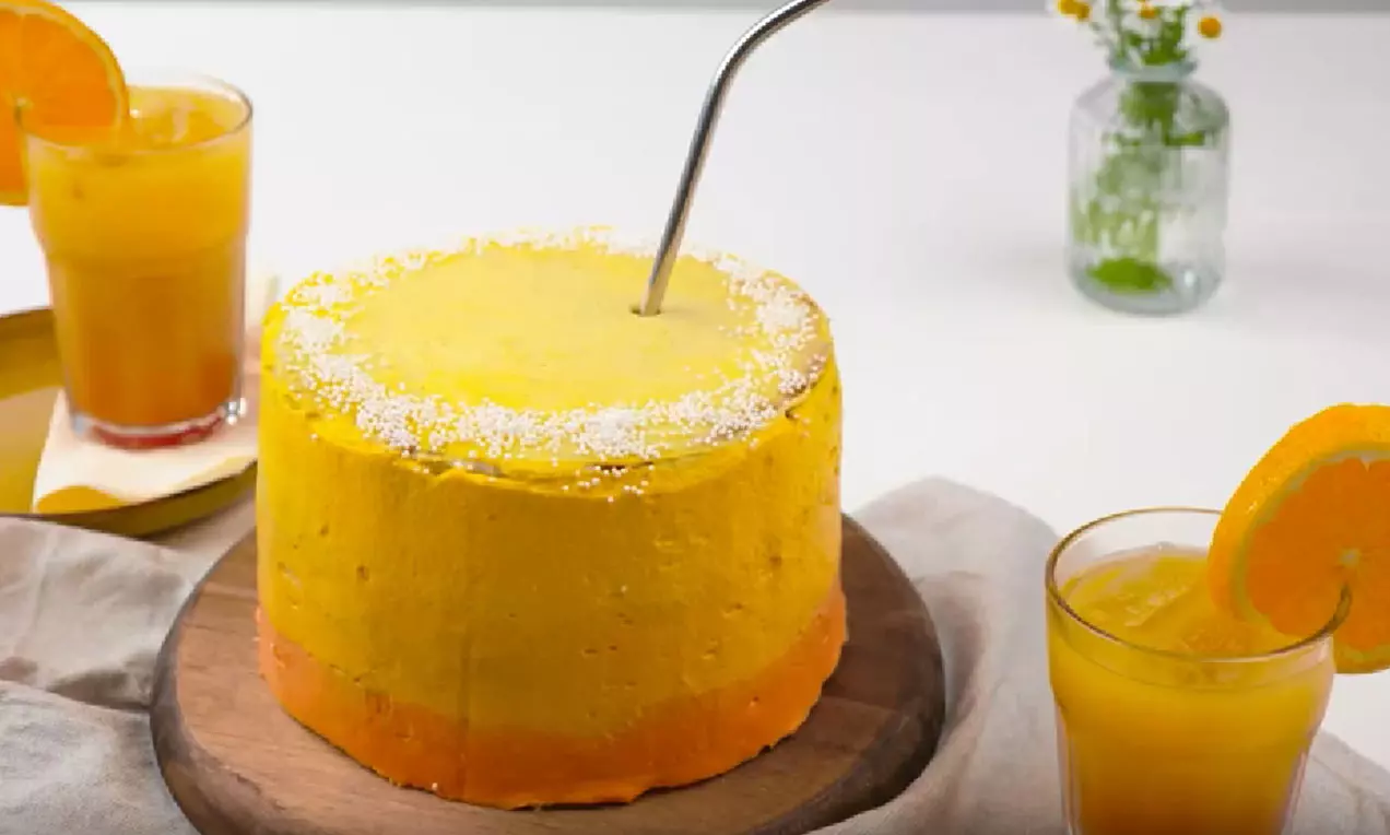 Gâteau cylindrique aux teintes dégradées d'orange et jaune vif, bordé de sucre en poudre, posé sur une planche en bois avec des verres de cocktail