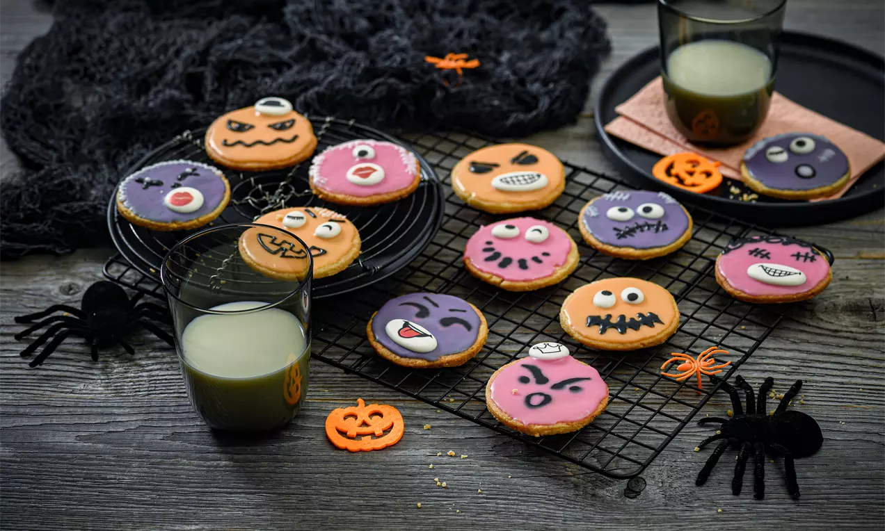 Bunte runde Halloween-Plätzchen mit gruseligen Gesichtern aus Zuckerguss auf schwarzem Kuchengitter, dekoriert mit Spinnen und Kürbisfiguren