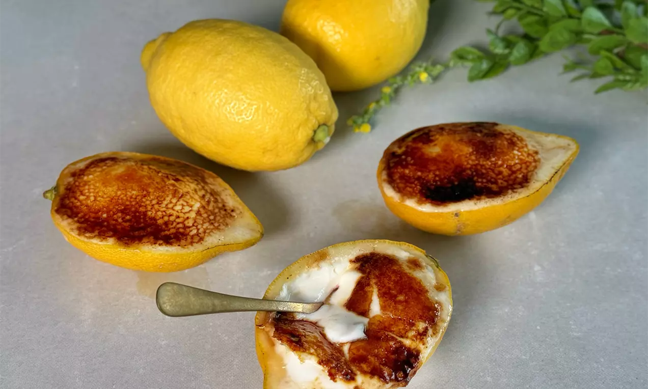 Hälften von Zitronenschalen mit goldbrauner karamellisierter Cremefüllung, daneben ganze Zitronen und ein grüner Zweig auf hellem Untergrund