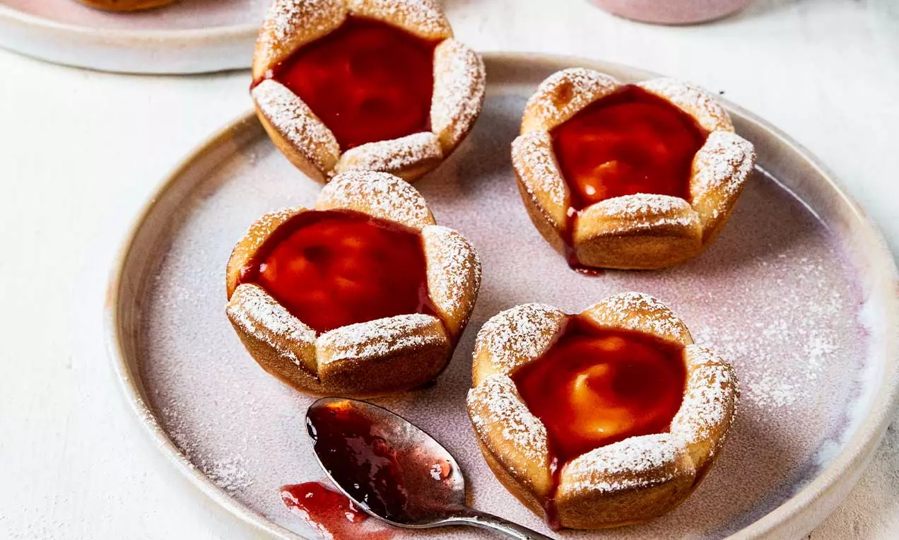 Petites pâtisseries dorées en forme de fleurs avec un cœur brillant de coulis rouge, saupoudrées de sucre glace sur une assiette beige