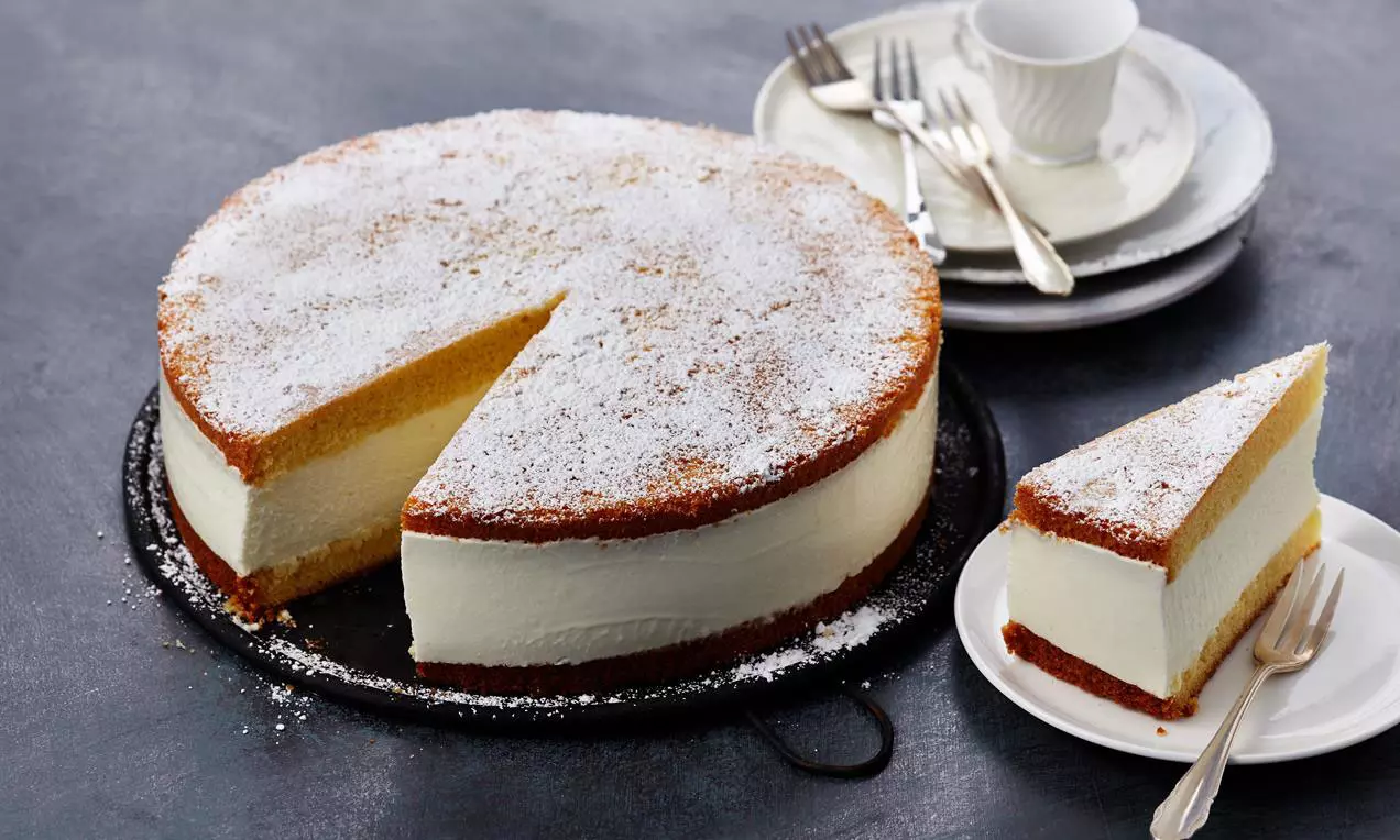 Käse-Rahm-Torte mit goldbraunem Boden und Deckel, weißer cremiger Füllung und Puderzucker auf schwarzem Teller, daneben Stück auf weißem Teller