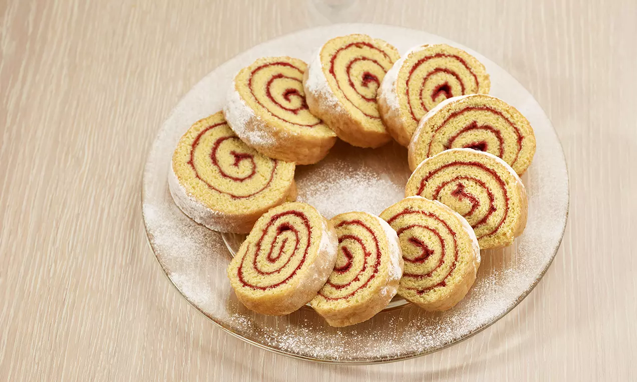 Fette di rotolo soffice dorato con spirali di marmellata rosso intenso, disposte in cerchio su un piatto cosparso di zucchero a velo