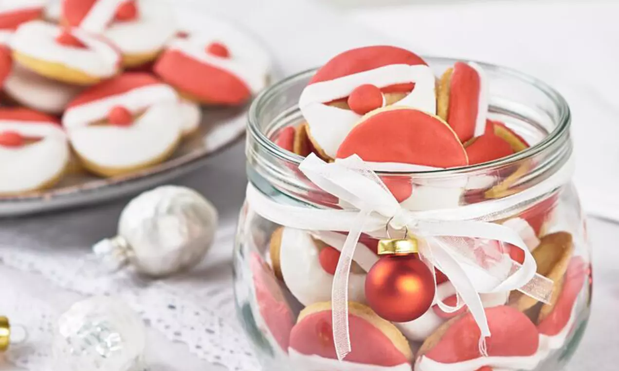 Petits biscuits colorés en rouge et blanc en forme de têtes de Père Noël dans un bocal en verre décoré d’un ruban et boule rouge