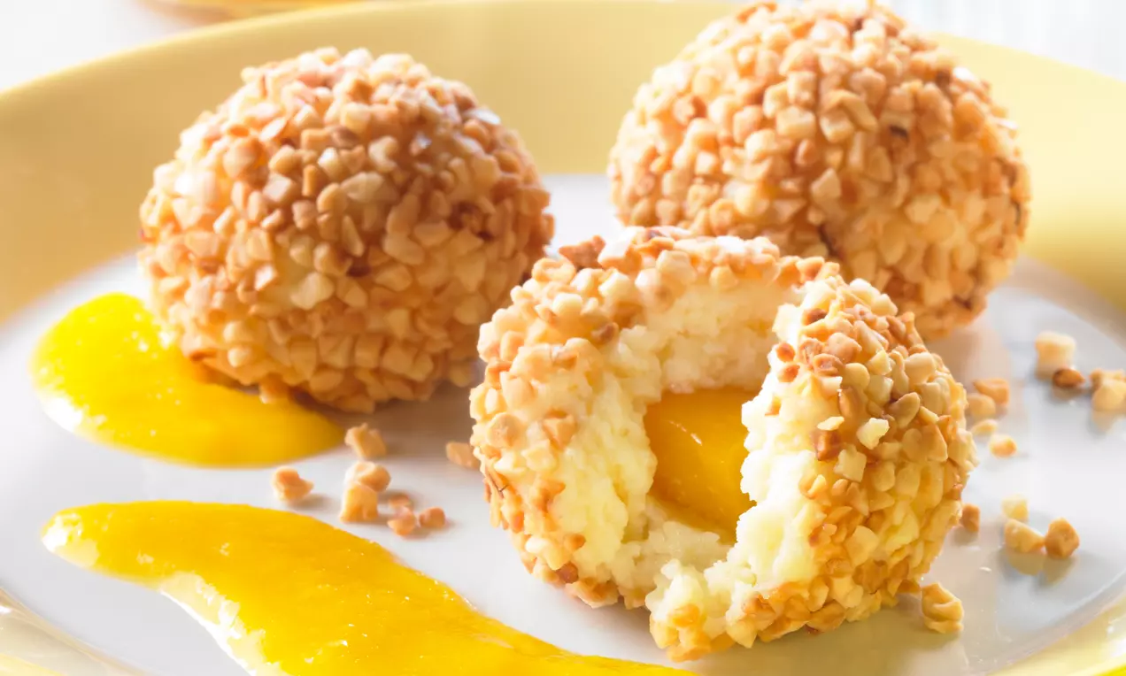 Boules dorées aux éclats de noisette sur une assiette beige, garnies d’un cœur de pêche juteuse avec un coulis jaune vif