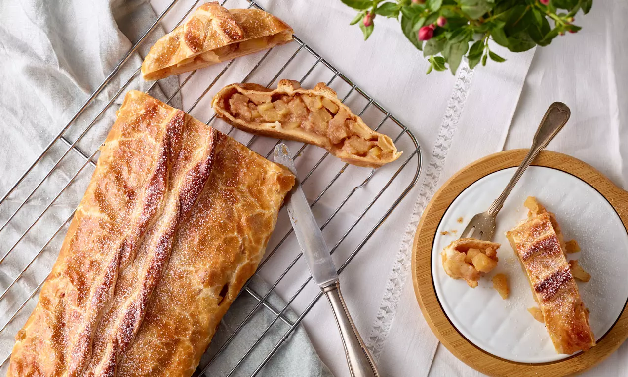 Apfelstrudel dorato con sfoglia croccante e ripieno di mele morbide, disposto su una griglia accanto a un piatto bianco decorato