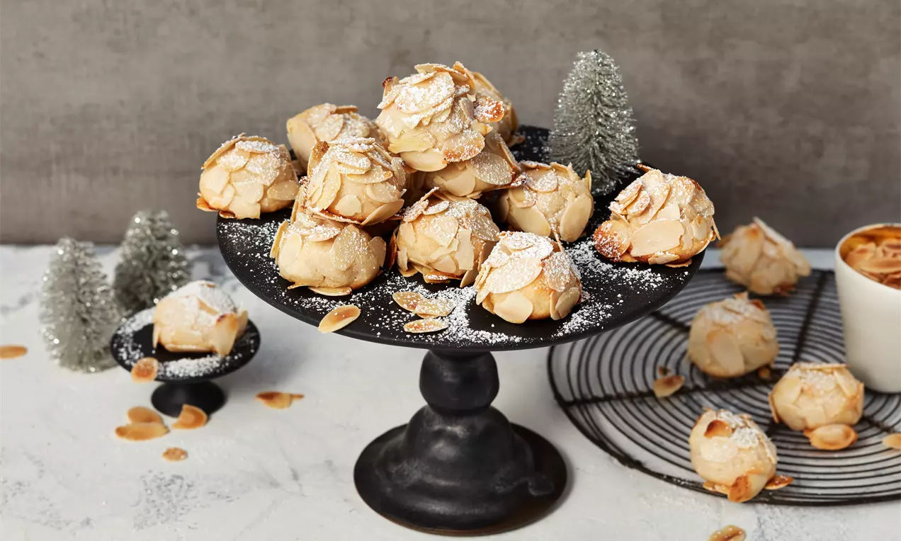 Marzipan-Wölkchen mit leicht gebräunten Mandelblättchen und Puderzucker auf schwarzer Etagere, umgeben von Miniatur-Tannen und Mandelsplittern