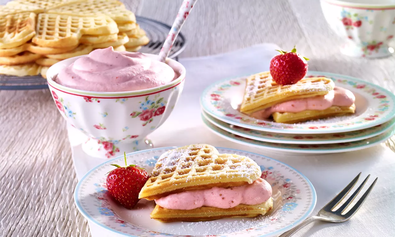 Goldgelbe, herzförmige Waffeln mit rosa Erdbeercremefüllung, dekoriert mit frischen Erdbeeren, auf floralen Tellern und hellem Stoff serviert
