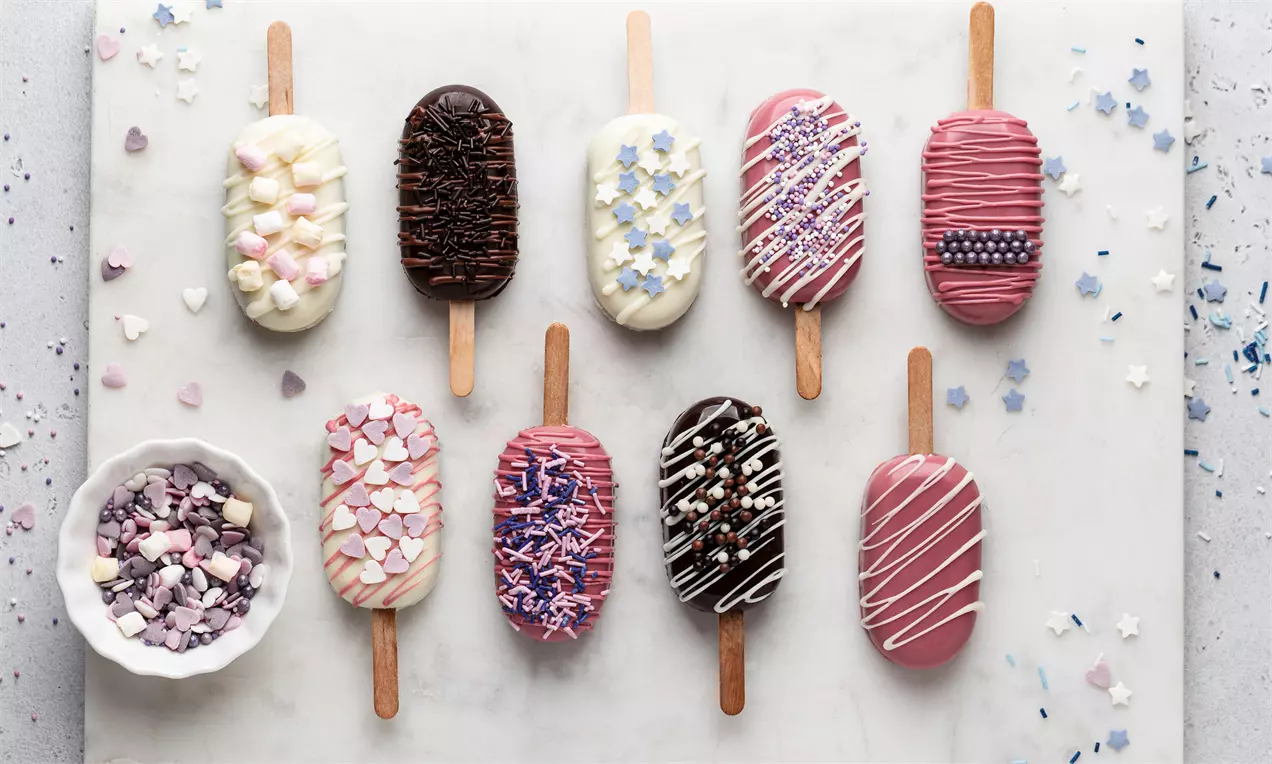 Mini gâteaux glacés en bâtonnets aux couleurs rose, blanc et chocolat décorés de perles, vermicelles ou éclats colorés sur un plateau marbré