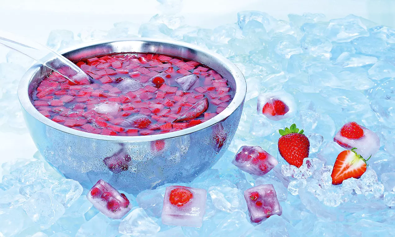 Metallschale mit roter Rhabarber-Bowle, darin Früchte und Eiswürfel, umgeben von losem Eis und Erdbeeren auf hellem Untergrund