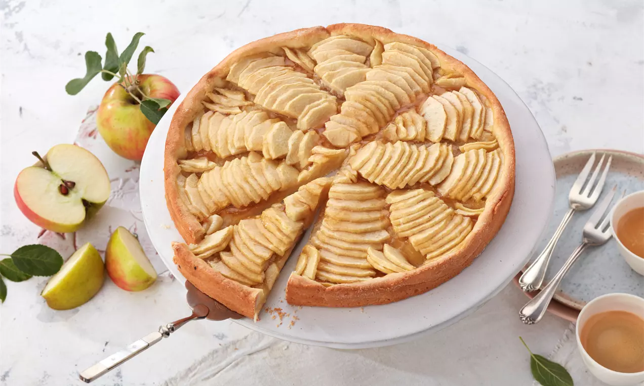 Apfeltarte mit goldgelbem Mürbeteig, belegt mit fächerförmig angeordneten Apfelscheiben auf einer weißen Kuchenplatte mit Kaffeeservice