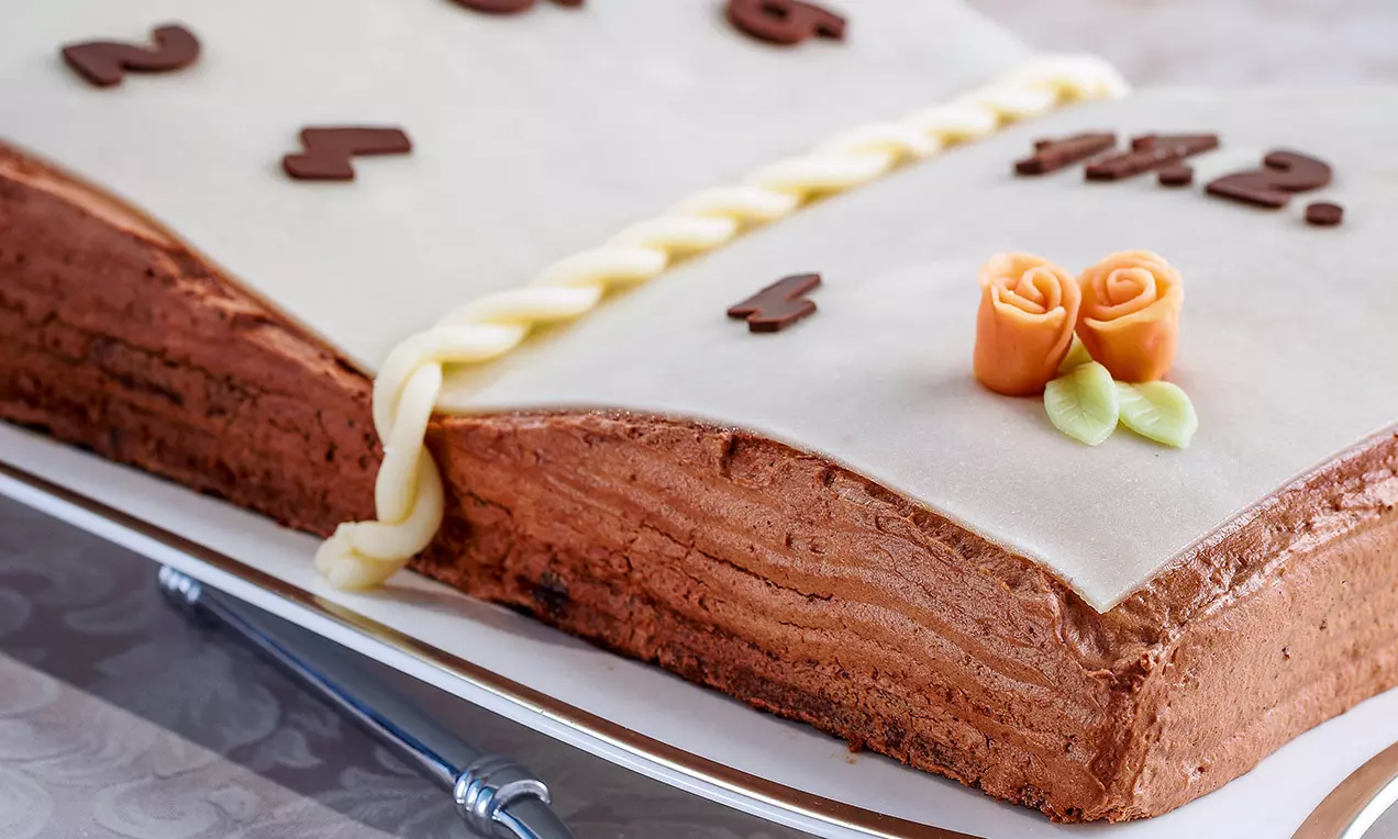 Torta a forma di libro aperto con copertura in pasta di zucchero bianca, decorata con numeri di cioccolato, roselline arancioni e foglie verdi