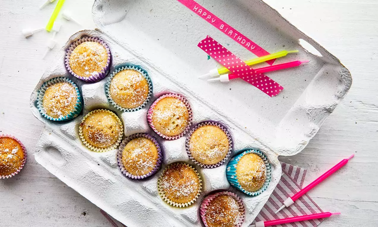 Petits muffins dorés saupoudrés de sucre dans des caissettes colorées disposés dans une boîte à oeufs blanche avec des bougies anniversaire