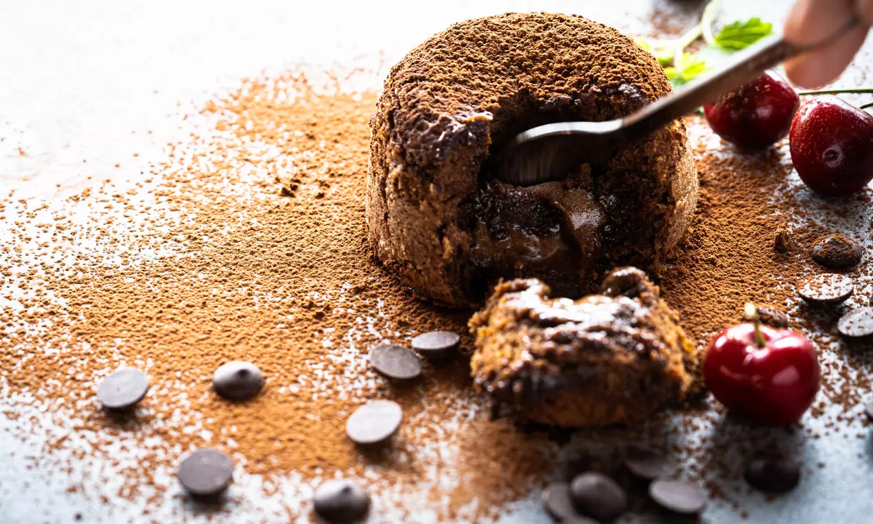 Schokoladenkuchen mit flüssigem Kern, bestäubt mit Kakao, angerichtet mit Kirschen, Schokotropfen und einem Löffel, der den Kern enthüllt