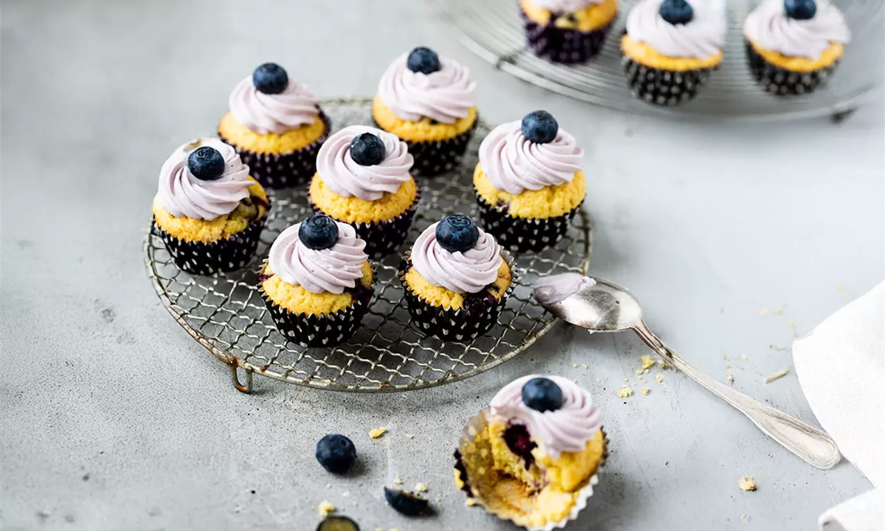 Petits muffins dorés garnis de crème violette tourbillonnée, décorés de myrtilles fraîches, disposés sur une grille métallique.