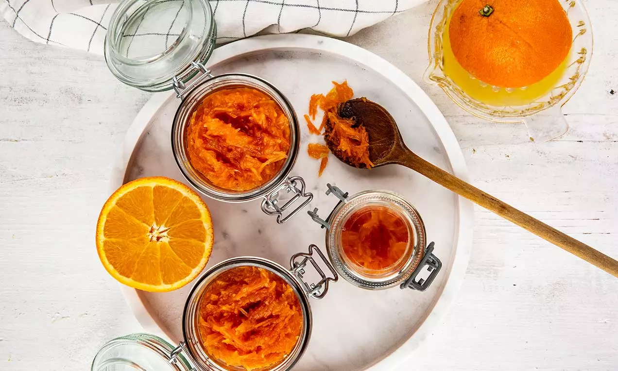 Confiture orange vif à la texture onctueuse dans des pots en verre, accompagnée d'oranges fraîches et d'une cuillère en bois sur un plateau blanc