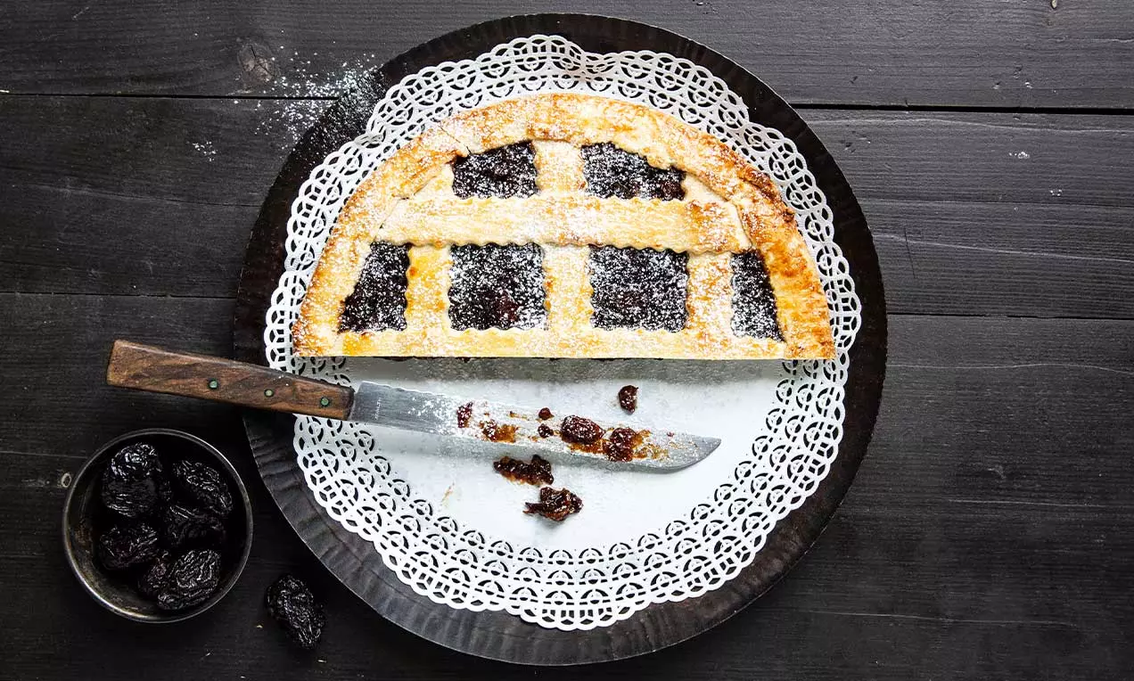 Crostata dorata con griglia di pasta friabile, ripieno scuro e lucido di prugne su centrino decorativo bianco e fondo nero