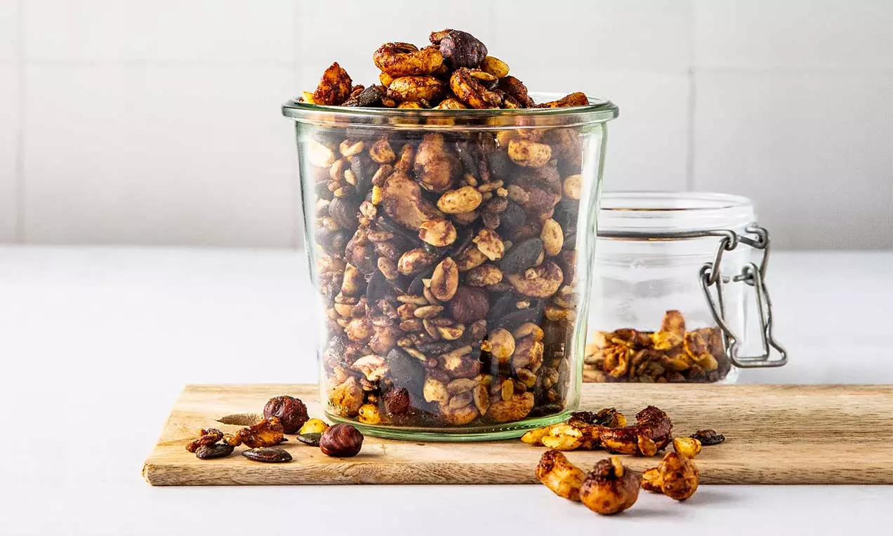 Mix croccante di noci dorate, arachidi e semi glassati al caramello leggero, disposto in un vaso di vetro su tagliere di legno chiaro