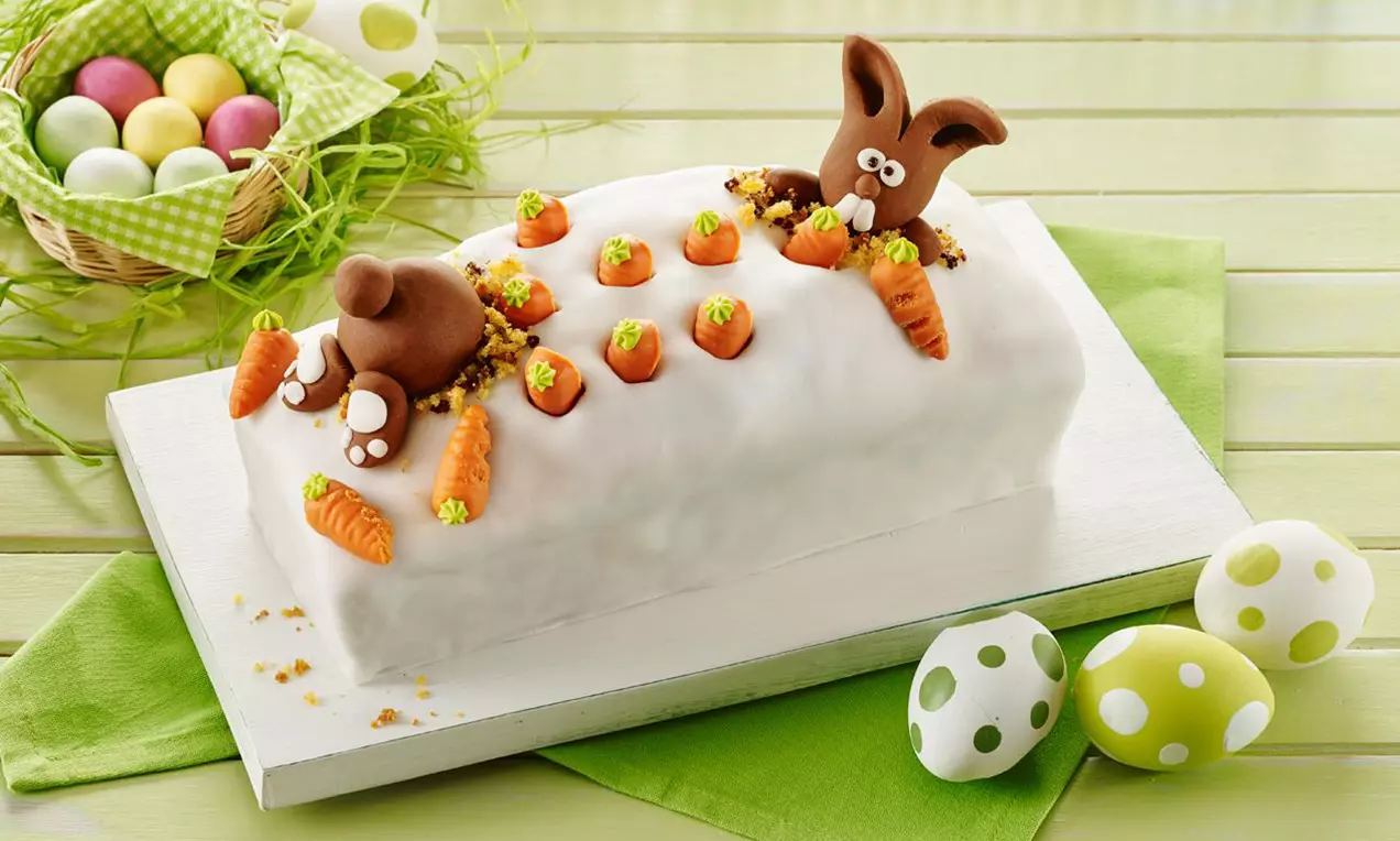 Torta rettangolare ripiena marmorizzata, ricoperta di glassa bianca decorata con carote e coniglietti di fondant su fondo verde e uova colorate