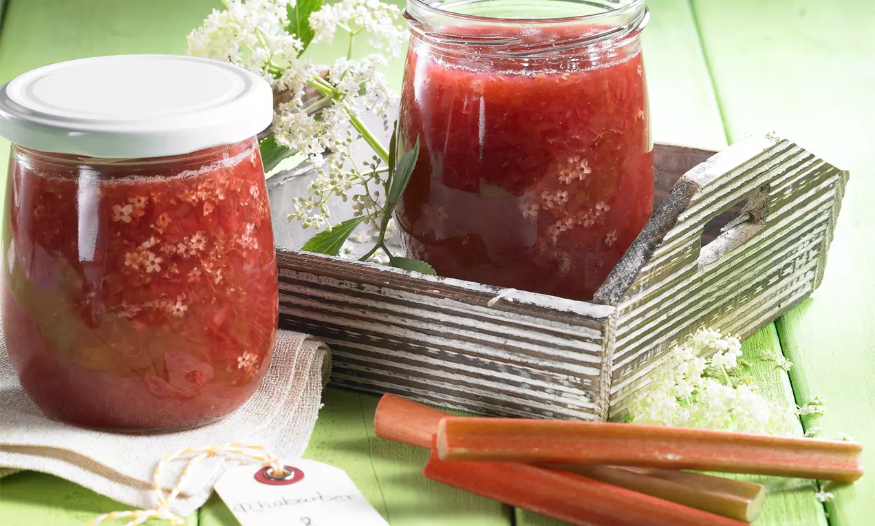 Rote Rhabarberkonfitüre mit feiner Textur in zwei Gläsern, dekoriert mit zarten weißen Holunderblüten auf grünem Holzlattenuntergrund