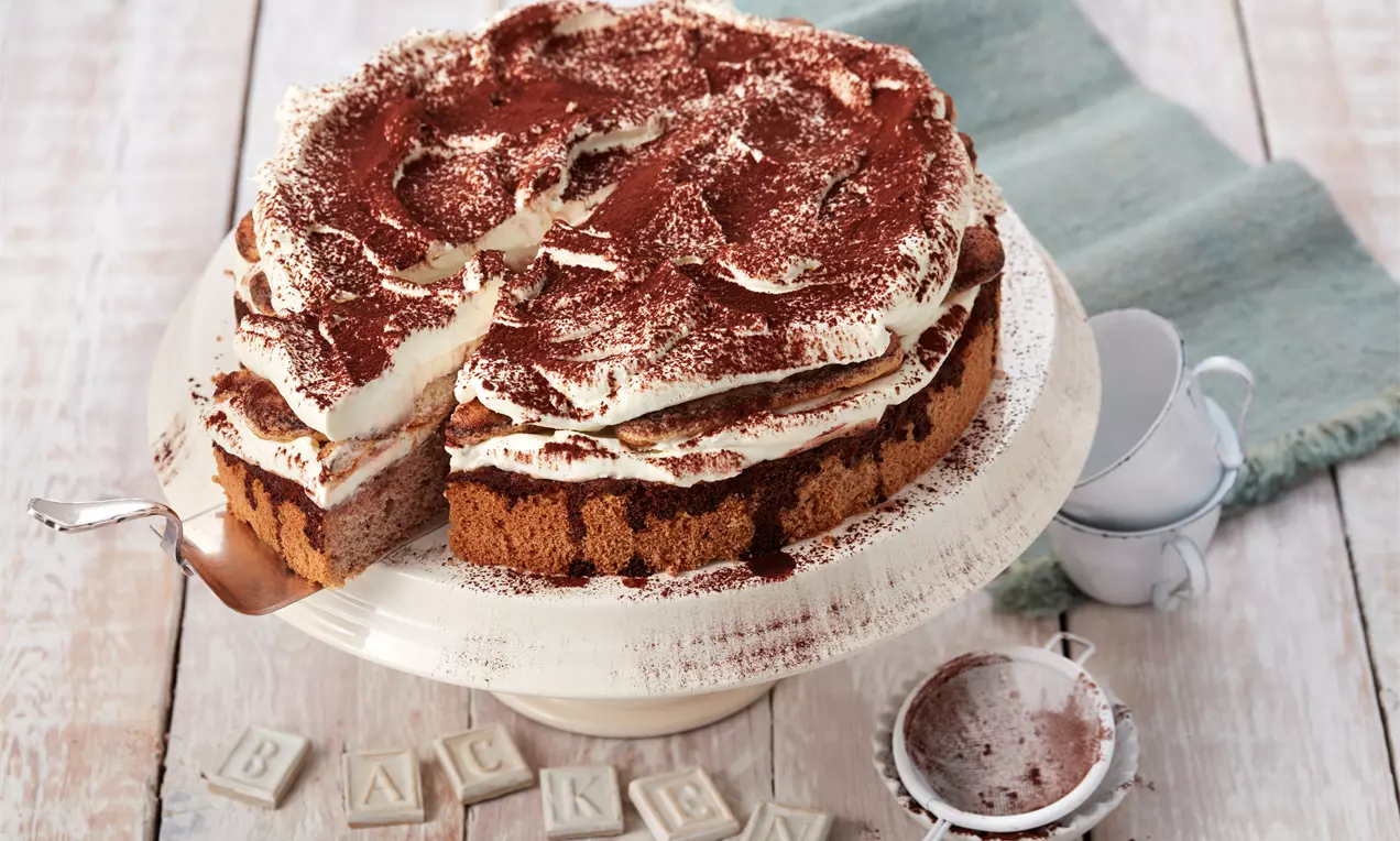 Tiramisu-Torte mit hellbraunem Löffelbiskuitboden, geschichteter Creme und Kakao bestäubt auf einer weißen Tortenplatte mit Sieb und kleinen Tassen