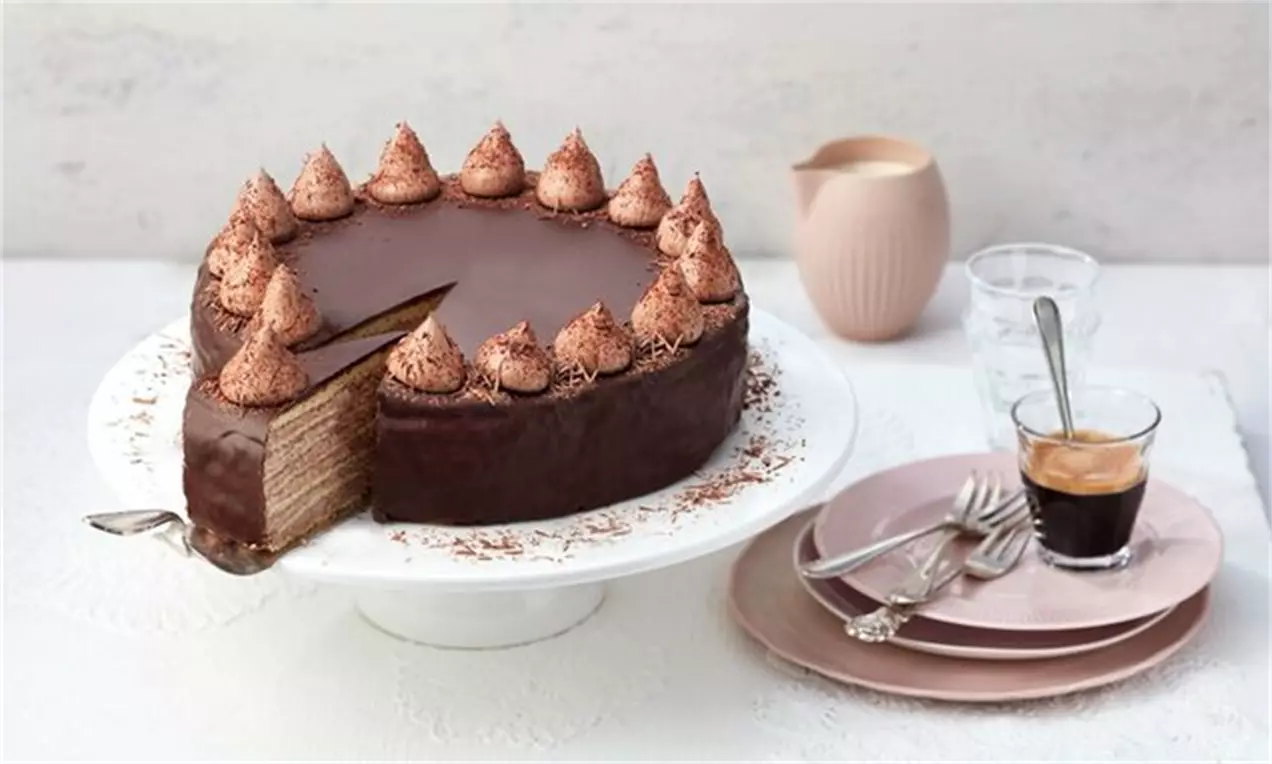 Schichttorte mit glänzender Schokoladenglasur, dekoriert mit Kakaocremehäubchen, angeschnitten auf weißer Tortenplatte mit Espresso und Teller