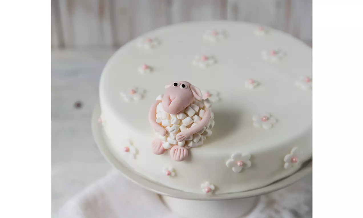 Ein rosa Schäfchen aus Fondant mit weißer Fellstruktur sitzt mittig auf einer schokoladenbraunen Torte mit weißen und rosafarbenen Blüten