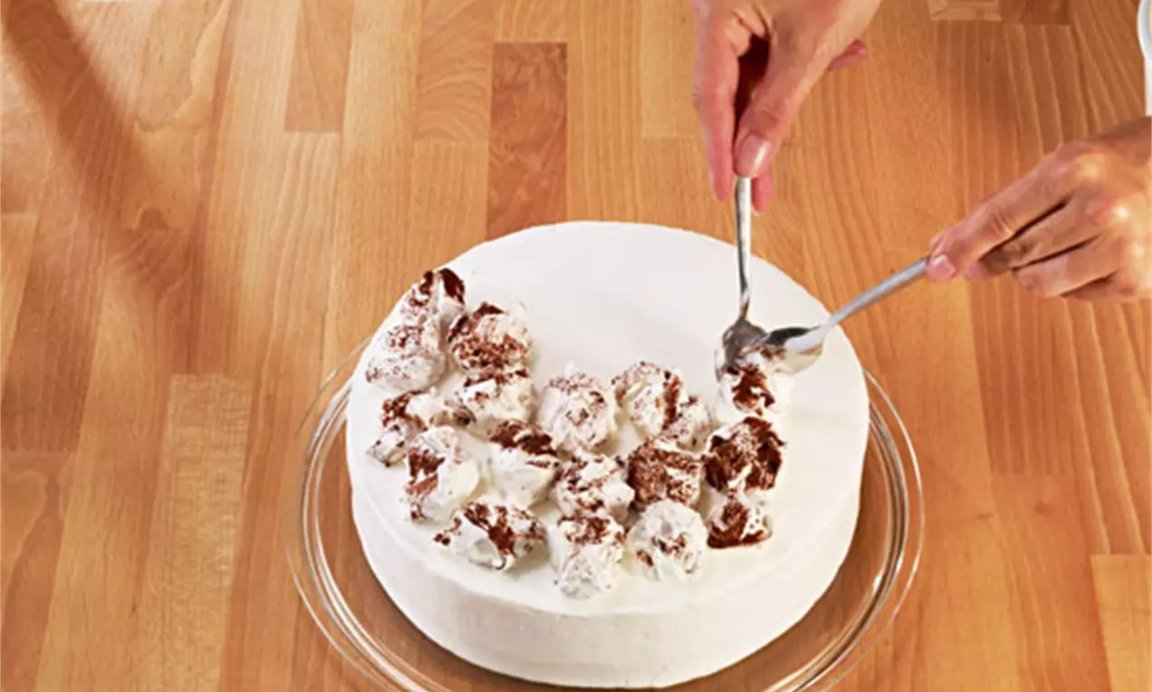 Eine weiße, runde Torte mit lockeren Sahnehäubchen, teils mit Kakao bestäubt, auf einer Glasplatte auf hellem Holztisch