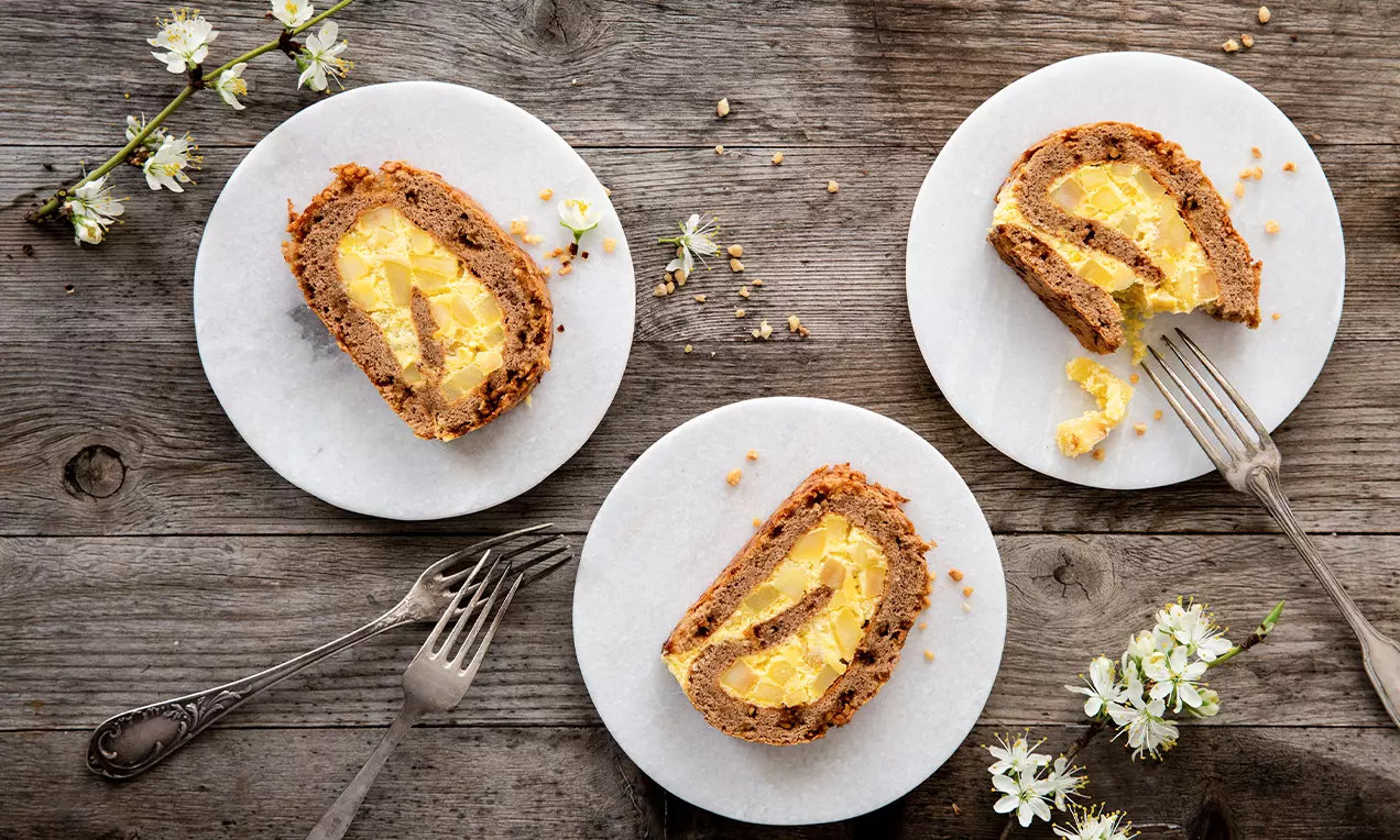 Tranches de gâteau roulé doré garnies de crème jaune et éclats de nougatine sur des assiettes blanches avec des fleurs de pommier