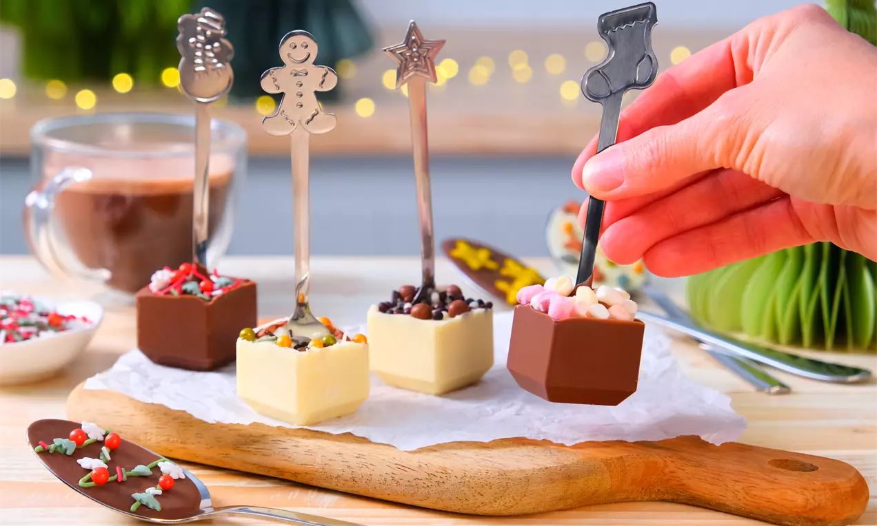Cuillères métalliques aux poignées festives plongées dans des cubes de chocolat blanc et brun garnis de mini pépites et guimauves