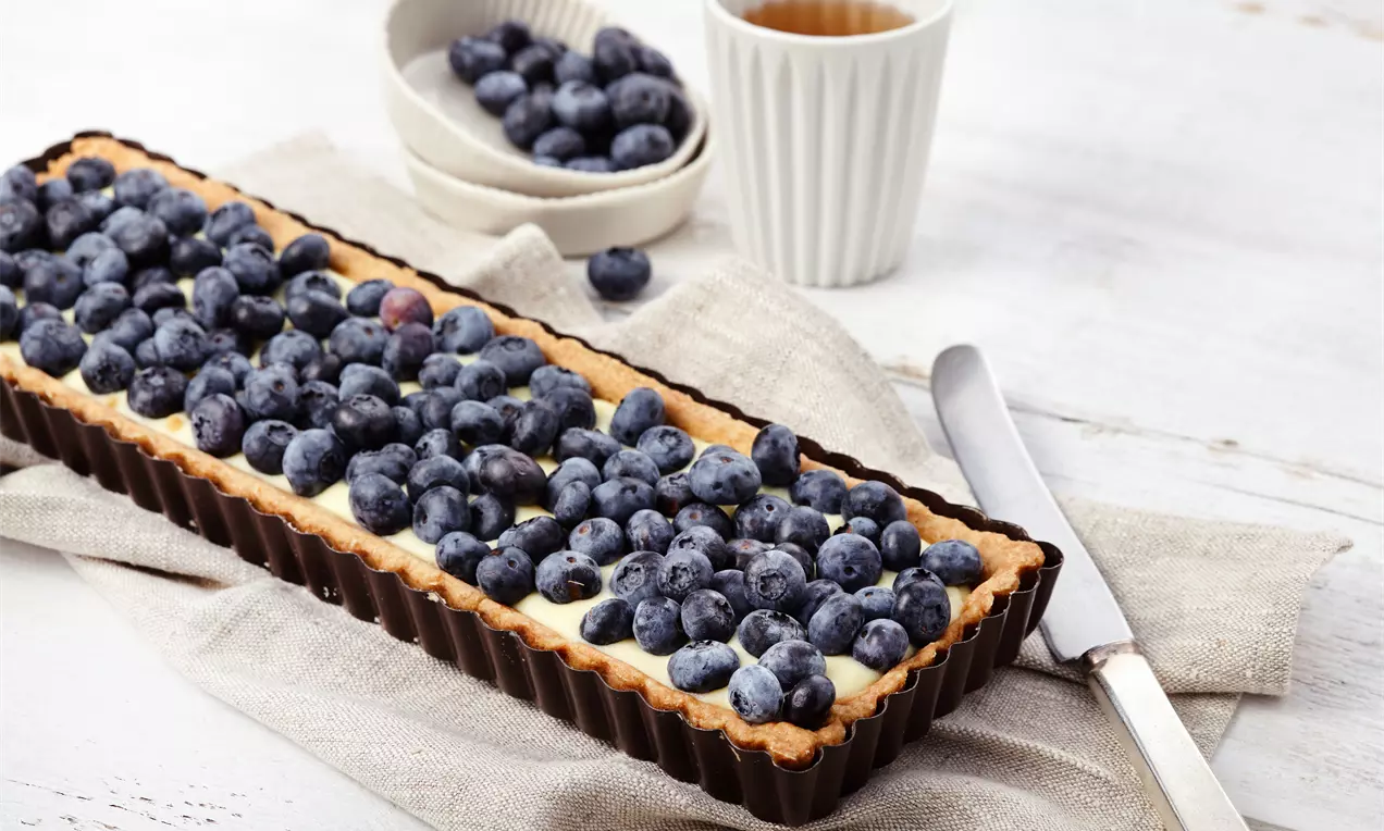 Rechteckige Heidelbeer-Tarte mit goldbraunem Rand und heller Crème Pâtisserie, dicht belegt mit saftigen blauen Heidelbeeren auf Leinentuch