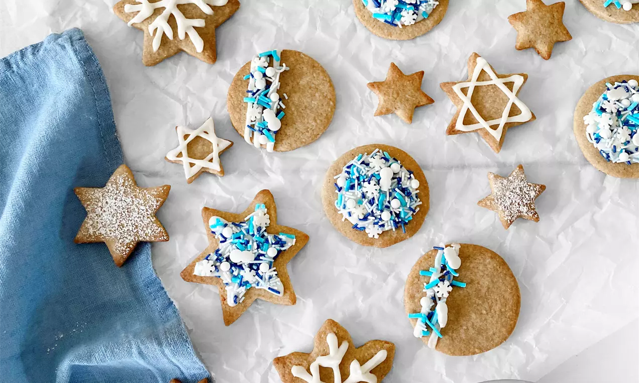 Biscotti dorati e friabili decorati con glasura bianca e zuccherini blu a forma di stelle e cerchi su carta bianca accanto a un panno celeste