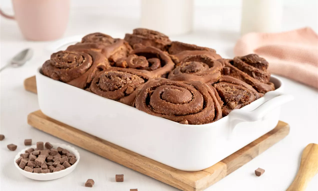 Schokoladen Zimtschnecken in weisser Gratinform auf einem Holzbrett mit rosa Mug und einem Porzellantopf im Hintergrund. Aufgnommen im Querformat