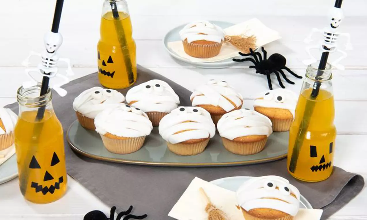 Cupcakes dorés enserrés de bandes de glaçage blanc strié, yeux en sucre, disposés sur un plateau gris avec araignées et jus orange