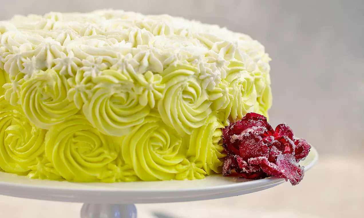 Torta decorata con volute di crema bianca e gialla, bordi dettagliati con rose di glassa e petali rosso intenso cosparsi di zucchero