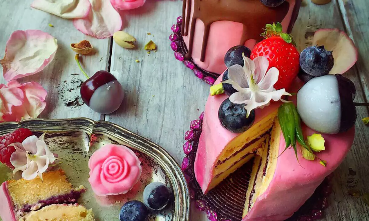 Ein kleines Törtchen mit rosafarbener Buttercreme, Blaubeeren, Erdbeere, weißen Blüten und Pistazien, aufgeschnitten auf rustikalem Holztablett