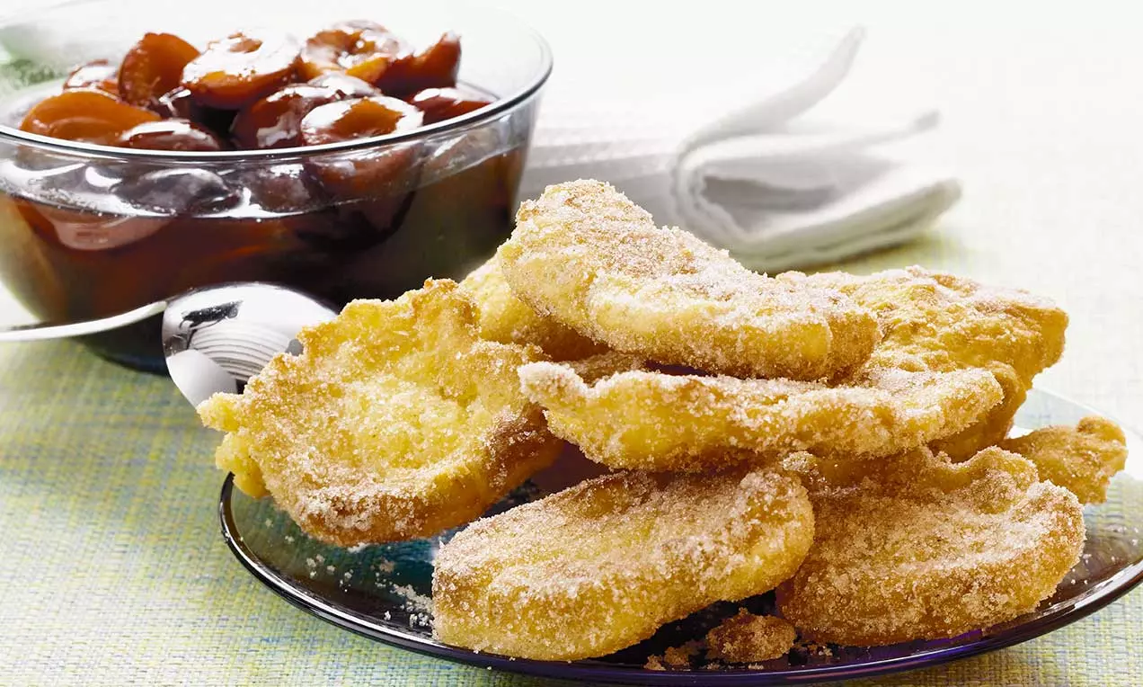 Beignets dorés et croustillants saupoudrés de sucre, disposés sur une assiette transparente avec un bol de compote brun foncé