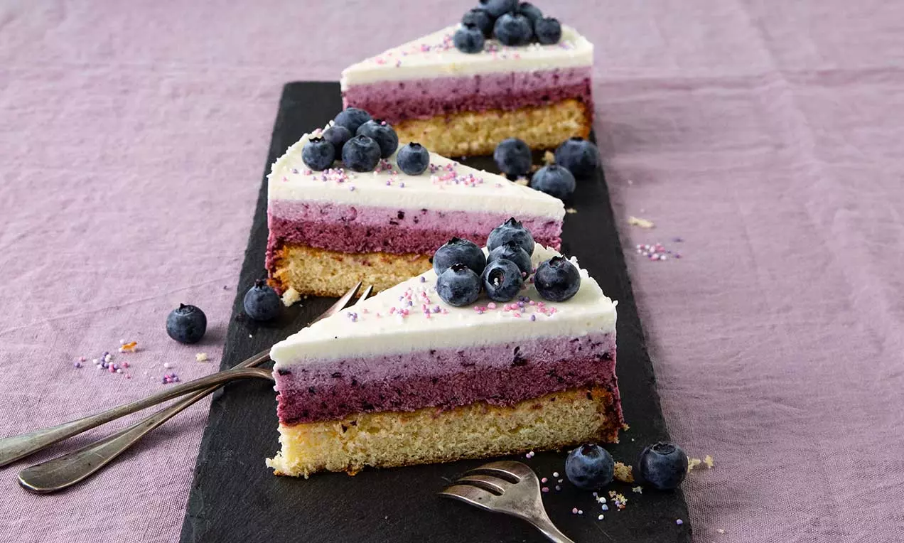 Fette di torta stratificata dai toni ombré bianchi e viola, con base dorata, guarnite con mirtilli freschi su un piatto scuro