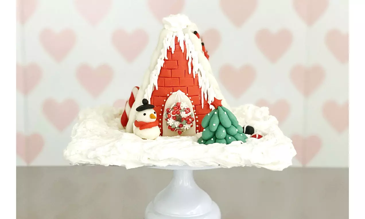 Knusperhäuschen aus roten Zuckermauersteinen mit weißer Zuckerglasur, dekoriert mit Schneemann, Baum und Süßigkeiten auf schneeweißem Untergrund
