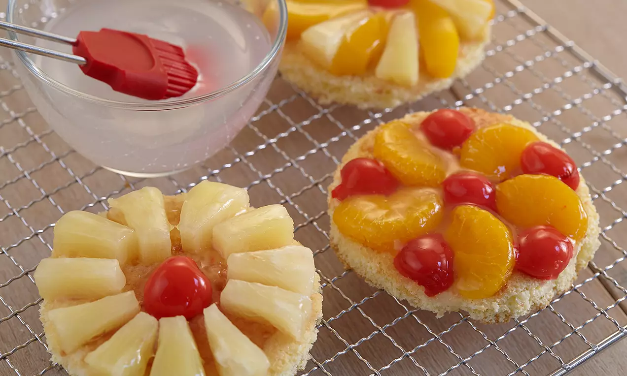Runde Fruchttörtchen mit glänzendem Belag aus gelben Pfirsichspalten, roten Kirschen und hellen Ananasstücken auf einem silbernen Kuchengitter