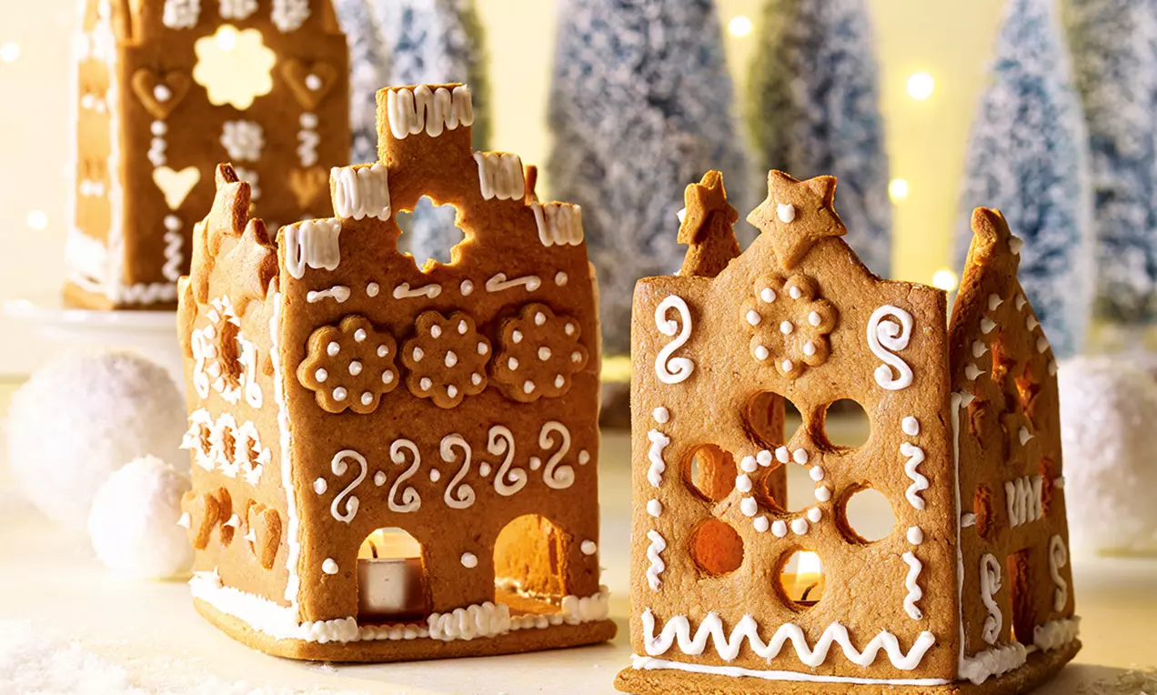 Petites maisons en pain d'épices brun décorées de glaçage blanc avec des ouvertures lumineuses, posées devant des sapins enneigés