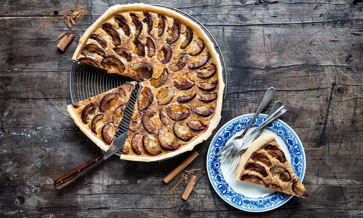 Torta rustica con fette di prugne scure disposte a spirale su un sottile strato dorato, servita con una fetta su piatto decorato blu e bianco