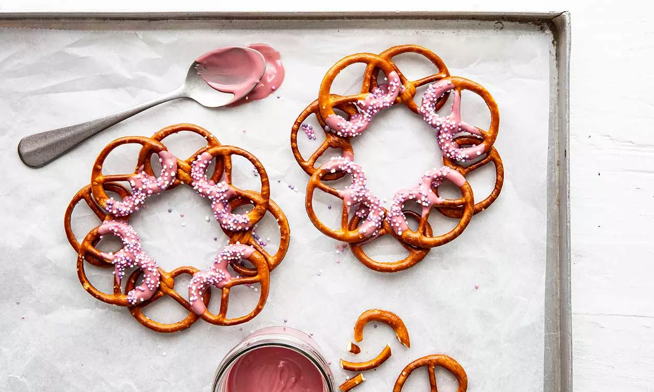 Couronnes de mini bretzels dorés assemblés en cercle, partiellement enrobées de glaçage rose et saupoudrées de perles sucrées blanches