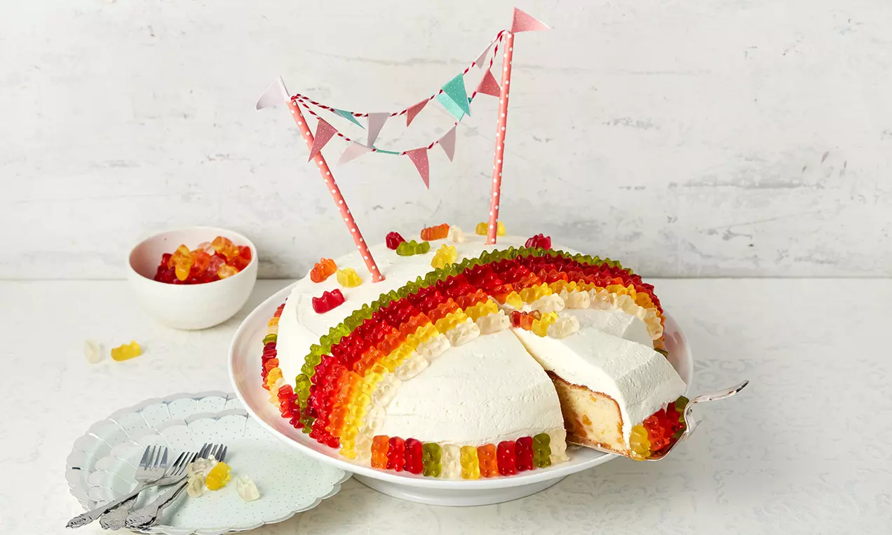 Halbkreisförmiger Sahnetorte mit Streifen aus roten, gelben und grünen Gummibärchen, dekoriert mit bunter Wimpelkette auf weißer Platte