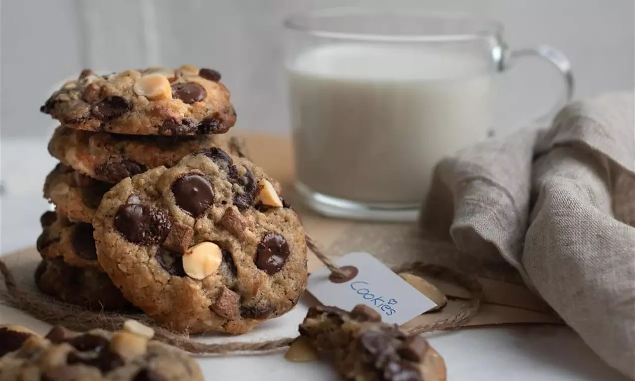 Vanille-Cookies mit goldbrauner Oberfläche, Paranussstücken und Schokodrops auf Holzbrett, daneben ein Glas Milch und beiges Stofftuch