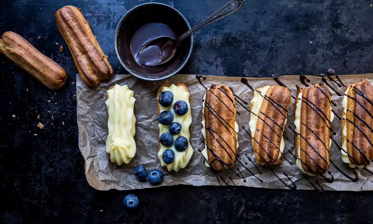 Goldbraune Eclairs mit heller Cremefüllung, dunklem Schokoladenguss, frischen Heidelbeeren und Schokoladensauce auf braunem Papier drapiert