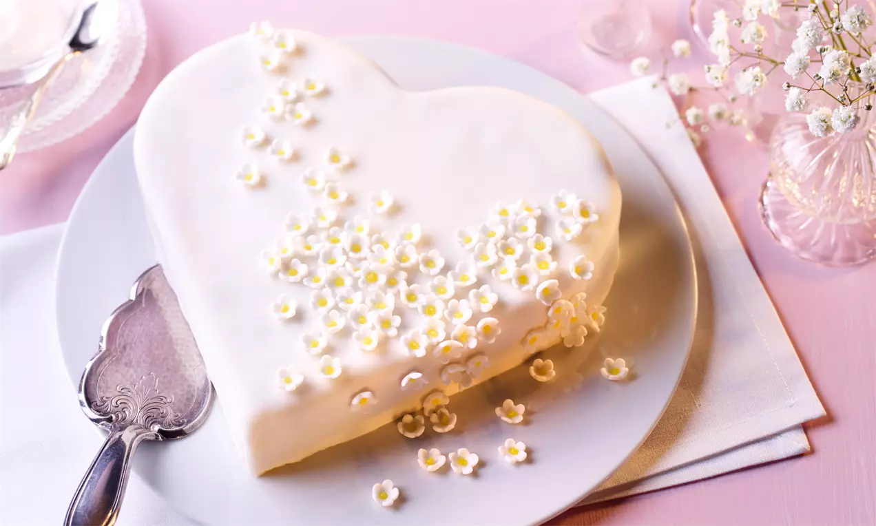 Herztorte mit weißer Glasur, verziert mit kleinen cremefarbenen Blüten, auf einem weißen Teller, neben Besteck und zarten Schleierkrautzweigen