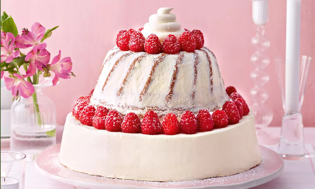 Zweistöckige Torte mit weißer Sahneschicht, zwei Himbeerkränzen und Sahnehäubchen, auf einer glitzernden Glasplatte mit rosa Hintergrund