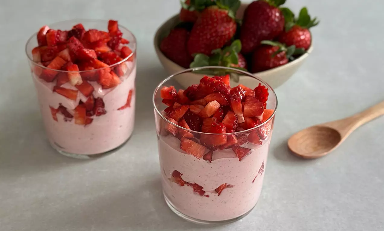 Zwei Gläser mit rosa Erdbeer-Quarkcreme, bedeckt mit glänzenden roten Erdbeerstückchen, daneben eine Schale frischer Erdbeeren und ein Holzlöffel
